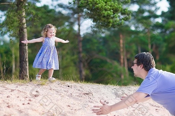 在一个阳光明媚、温暖的夏日，一个快乐的小卷发女孩和她的父亲玩耍，从松林中的沙丘上奔跑