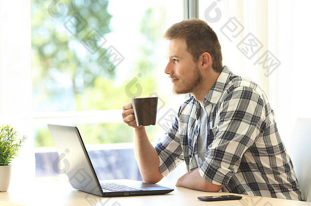 一个忧郁的男人拿着一台笔记本电脑从窗户往外看，手里拿着一个咖啡杯