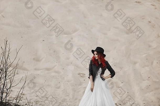 穿着皮夹克、黑帽子和<strong>时尚婚纱</strong>的美丽时尚红发模特儿在沙丘前摆姿势