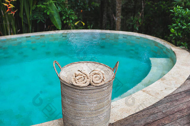 两条米色卷毛巾放在柳条篮子里，靠近私人游泳池，池水清澈，呈青绿色