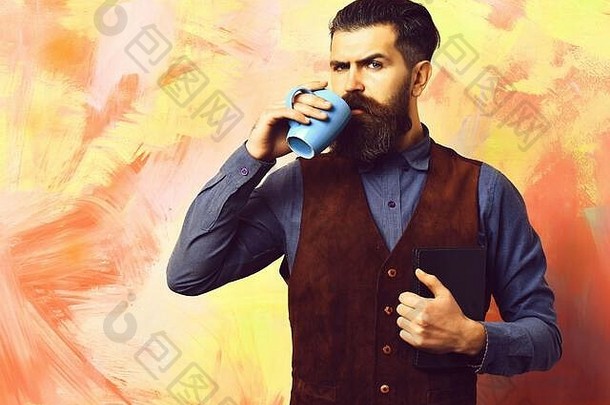 有胡子的男人。长胡子残酷的高加索人赶时髦的人胡子持有蓝色的咖啡杯黑色的笔记本古董衬衫皮革背心色彩斑斓的工作室背景