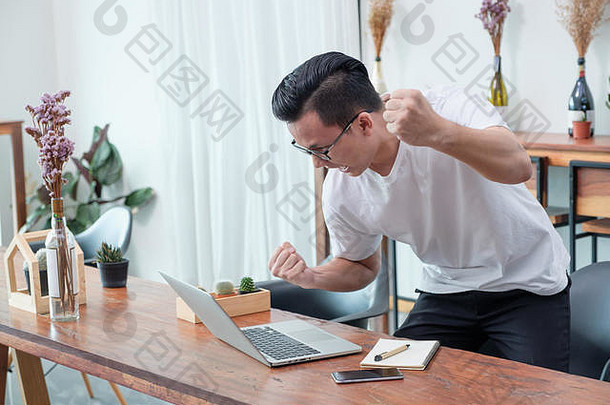 亚洲休闲男士站在咖啡厅的笔记本电脑旁，用手机庆祝好消息，成功的商业理念，在办公室外工作，在家里工作