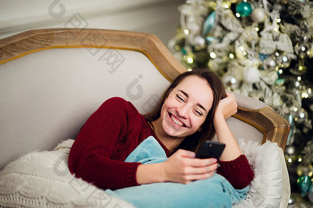 年轻的女人检查文本消息移动电话liyng沙发前面圣诞节树首页