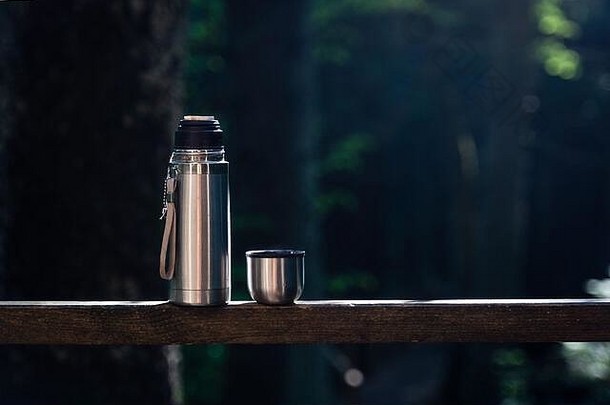 阳台上的木栏杆上有一个装有茶叶的金属保温瓶和一个杯盖，可以俯瞰森林。