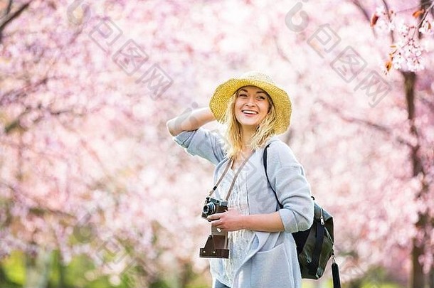 戴草帽的美女在樱花盛开的美丽公园里旅行，用复古相机拍照。背包游客