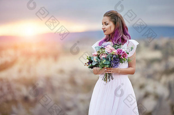 在黎明和日落的余晖中，手里拿着一束美丽鲜花的女人站在山上。漂亮的白色长裙穿在女孩b身上
