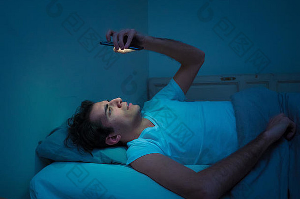 瘾君子深夜在床上用智能手机聊天和上网。在光线阴暗的房间里无聊、失眠、疲倦。在里面