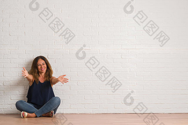 中年西班牙裔妇女坐在白砖墙上方的地板上，看着摄像机，张开双臂微笑着拥抱。愉快的表情
