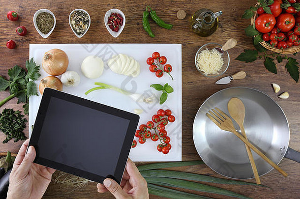 食物俯视图，手拿着数字平板<strong>电</strong>脑放在厨房的白色切菜板上，木制顶部有西红柿、蔬菜和<strong>炖锅</strong>