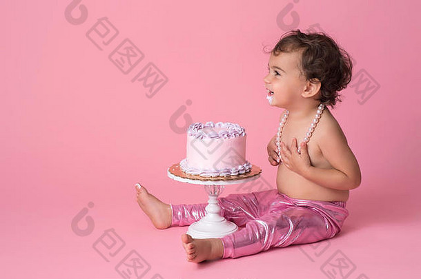 一个快乐的，一岁的，带着蛋糕坐着的小女孩。她穿着粉红色的紧身裤，一串珍珠，下巴上有霜。