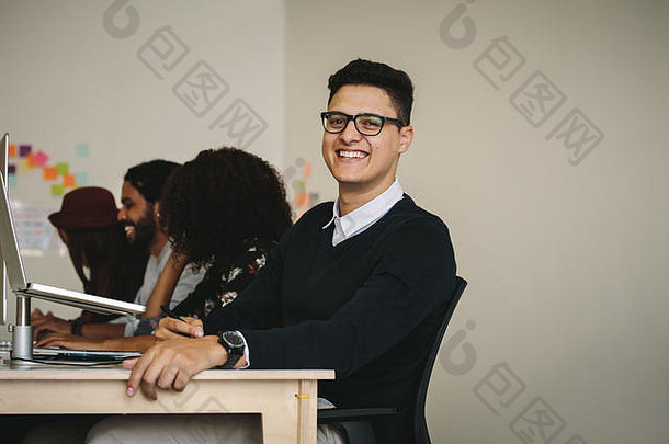 一位面带微笑的商人坐在办公桌旁，桌上放着一台笔记本电脑。企业家和他的同事坐在办公室里工作。