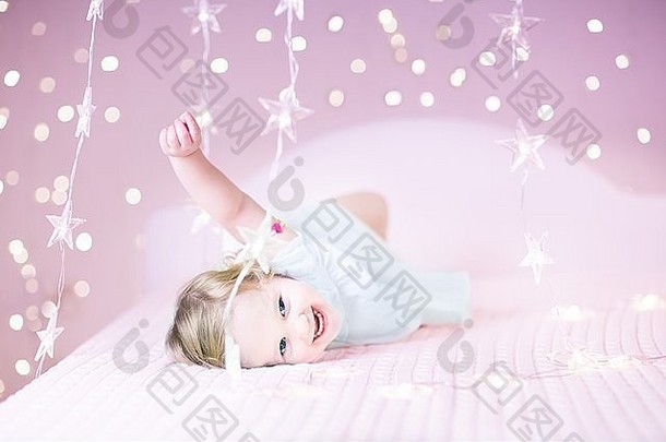 快乐笑蹒跚学步的女孩玩床上美丽的软粉红色的灯