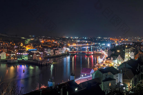 惠特比镇和海港的一幕在夜间亮了起来。明亮的五颜六色的灯光反射在水面上，头顶是一片漆黑的天空。
