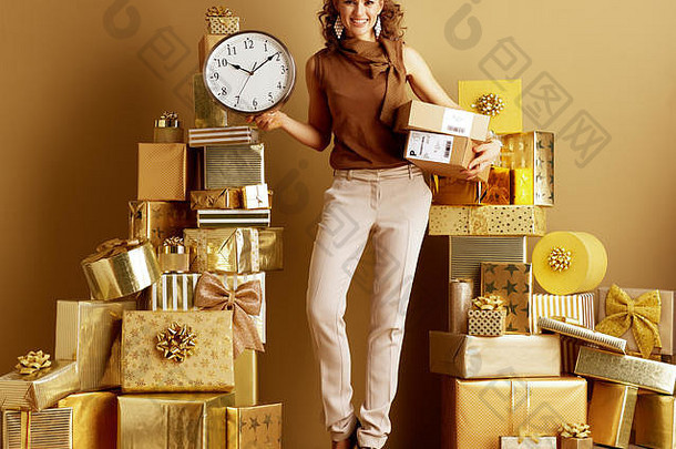 完整的长度肖像快乐现代女人黄金米色裤子棕色（的）上衣包裹显示时钟桩金礼物前面