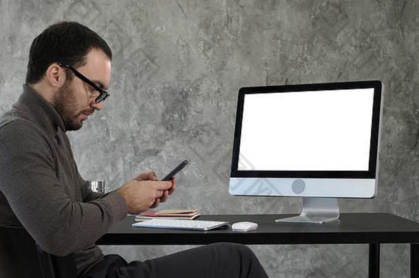 这位商人看着他的智能<strong>手机</strong>信息，坐在<strong>电脑</strong>屏幕旁。白色显示器。