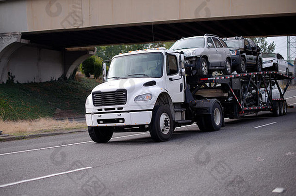 一辆白色中型汽车运输车半卡车在一座过桥下的公路上用一辆特殊的双层半挂车运输车辆