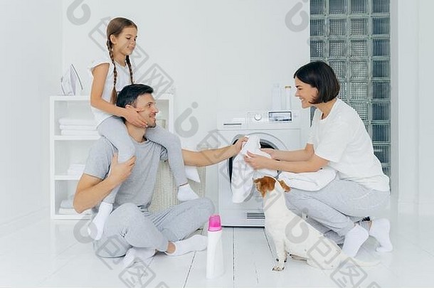 快乐的父亲把小猪还给女儿，帮妻子装洗衣机，他们的宠物在旁边摆姿势，白色地板上放着一瓶洗衣粉。<strong>友爱</strong>的