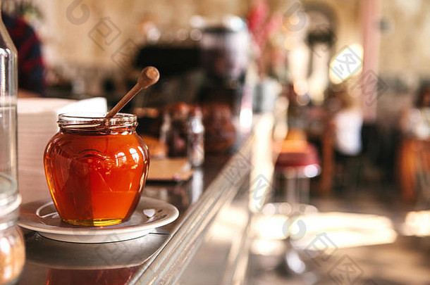 在一家背景模糊的咖啡馆里，美味的蜂蜜放在一个玻璃罐里，前景是一个特殊的木勺。