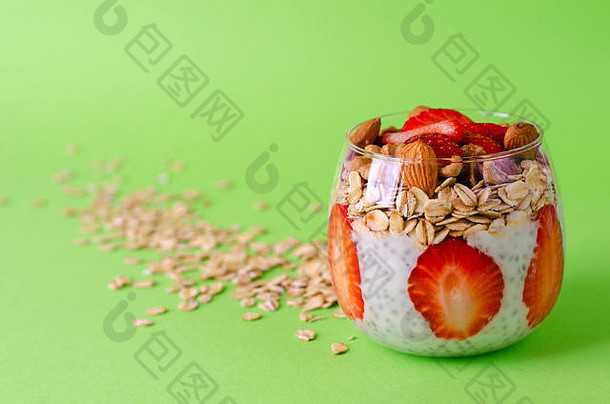 酸奶加布丁，配新鲜草莓、隔夜燕麦和坚果，置于绿色背景的玻璃杯中。前视图。均衡健康早餐理念。