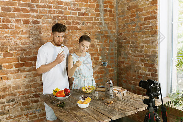 Vlogger和blogger自由职业，食品概念。一对年轻的高加索人夫妇在<strong>厨房</strong>的砖墙上用专业摄像机一起做饭，并为vlog和社交媒体录制现场视频。