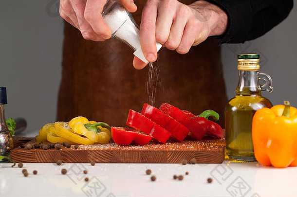 男厨师用盐瓶在甜椒中加盐。放在木制砧板上的蔬菜