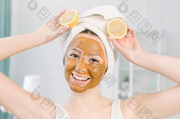 美容护肤理念。迷人的白种女人穿着白色毛巾和泥面膜，里面有两个半个柠檬，室内照在灯光下。