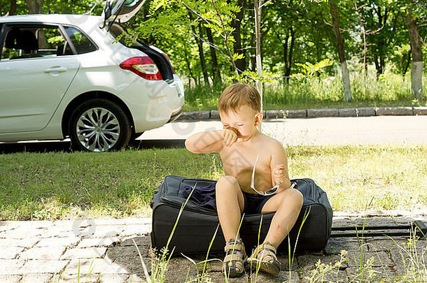 年轻的金发男孩坐在路边脱下衬衫的手推车旅行袋上打喷嚏