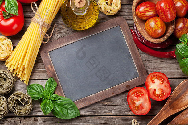 意大利食物烹饪。西红柿，罗勒，意大利面，橄榄油和黑板放在木制的餐桌上。前六名