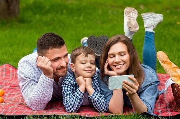 在公园野餐时使用智能手机观看娱乐视频的快乐家庭