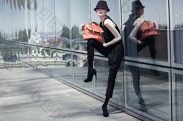 时尚的亚洲女孩，穿着黑色西装，戴着黑色帽子，摆着玻璃墙的姿势