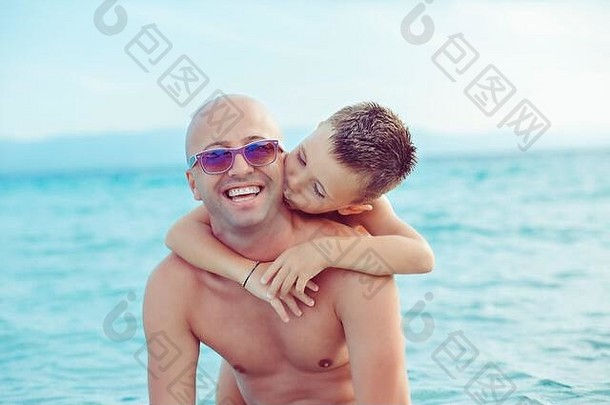 父亲戴着太阳镜，微笑着用双手拥抱儿子，而儿子则在户外背景蓝色的大海上亲吻他的脸颊