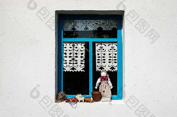 乡村农舍的白色墙壁，只有一扇蓝色的窗户，窗台上有一个布娃娃和花盆