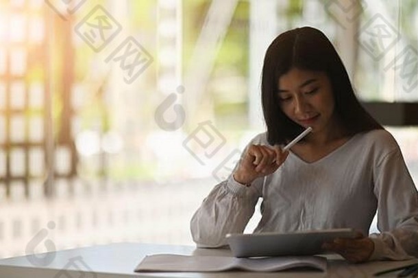 照片中，一位美丽的女士手里拿着一台电脑平板电脑和一支手写笔，坐在comfortabl的白色办公桌旁，看着它