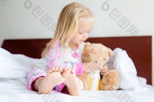 可爱的金发小女孩和玩具熊坐在床上。快乐的童年。在冠状病毒CVID-19大流行期间呆在家里