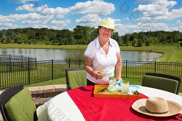 一位戴着时髦太阳帽、面带微笑的老太太站在室外露台上的花园餐桌旁，桌上盖着一副红布眼镜