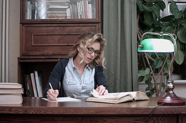 戴眼镜的年轻学生坐在桌子旁，从书上写笔记