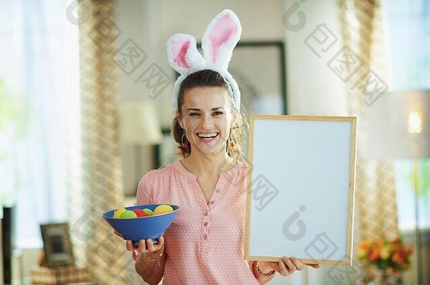快乐年家庭主妇粉红色的上衣复活节兔子耳朵蓝色的板色彩斑斓的复活节鸡蛋显示空白白色广告牌