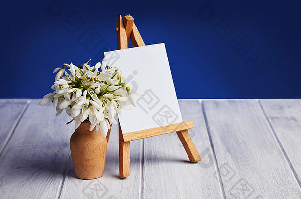 一束雪花莲和一个小画架，蓝色背景上有一张白纸和一个小罐子。带有文本空格的贺卡