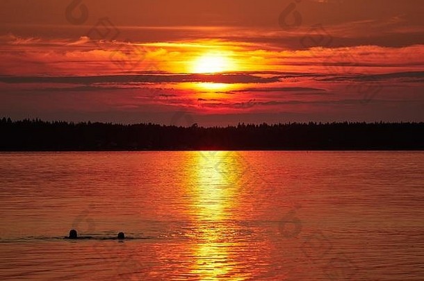在芬兰的瓦萨，红色的夕阳天空、平静的海水上的五彩缤纷的云彩和两个正在游泳的孩子。明亮的太阳圆盘部分被遮住了