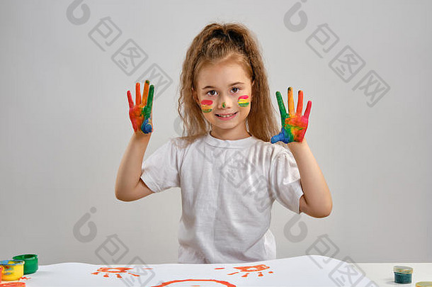 穿着白色t恤的小女孩站在桌旁，手里拿着惠特曼和五颜六色的颜料，展示着画过的手和脸。白色的隔离带。中等特写镜头。