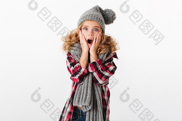 一个戴着冬季帽子和围巾的震惊小女孩看着隔离在白色背景下的摄像机的照片