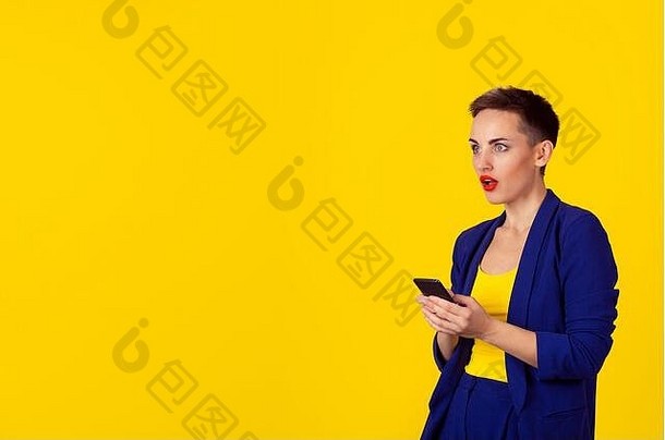 震惊的女商人拿着手机向旁边的空间望去，隔离了黄色背景墙。商务装正式蓝色套装，短发，红色