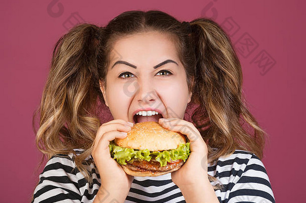 一个十几岁的女孩拿着牛肉汉堡三明治，脸上挂着快乐的表情，嘴里张着饥饿的嘴，背景是孤立的粉色。快餐概念。