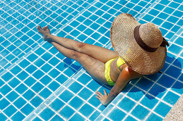 一位身穿黄色泳衣、戴着帽子和太阳镜的年轻女子站在<strong>游泳</strong>池里，在阳光明媚的<strong>夏日</strong>微笑着