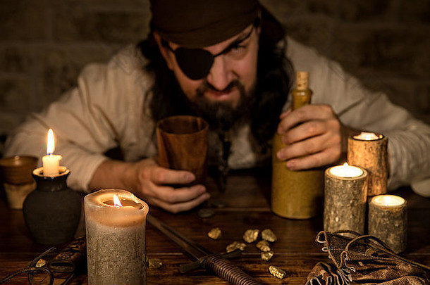 脾气暴躁的海盗拿着一瓶朗姆酒坐在一张中世纪的桌子上，桌上放着很多蜡烛