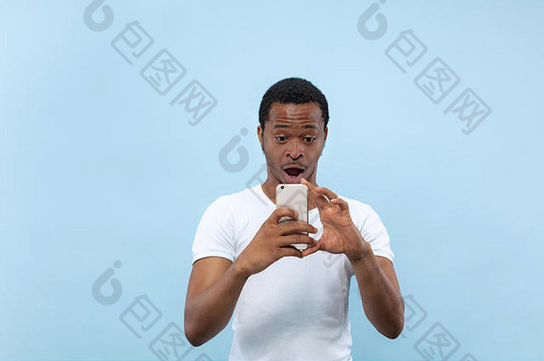 蓝色背景上穿着白衬衫的年轻非裔美国人的半身特写肖像。人类的情感，面部表情，广告概念。在他的智能手机上拍摄照片或视频日志内容。