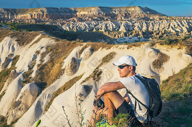 土耳其卡帕多西亚，一位戴着白色帽子、背着背包的年轻运动型男子坐在砂岩岩石的山顶上，观看自然景观。