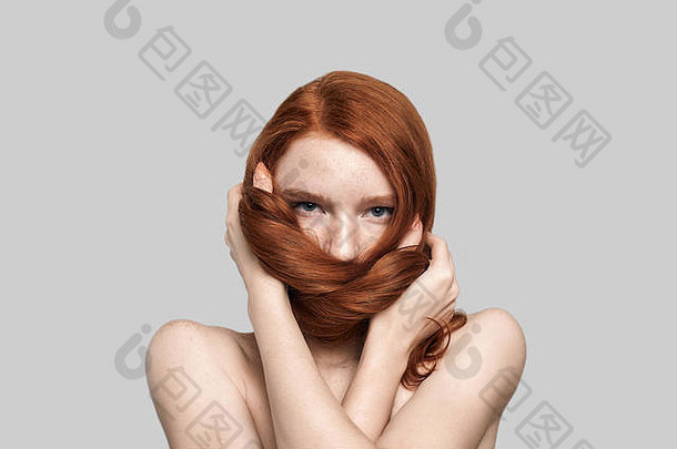 摄影棚拍摄了一位年轻漂亮的红发女子站在灰色背景下玩着她完美的长发。美女肖像