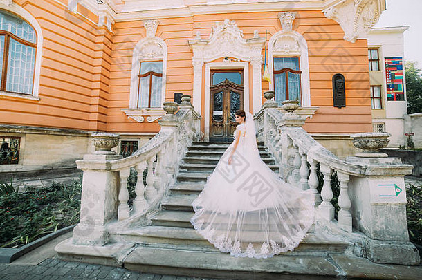 美丽的新娘穿着华丽的礼服，拖着长长的尾巴走上石头楼梯，来到浪漫的复古建筑