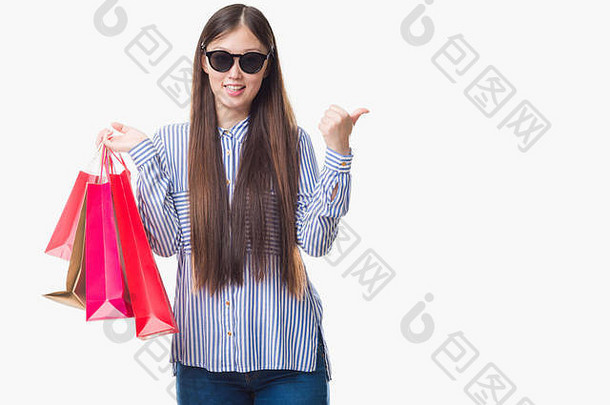 一位年轻的中国妇女站在孤立的背景下，手持购物袋，指着卖场，拇指朝上，脸上带着幸福的微笑